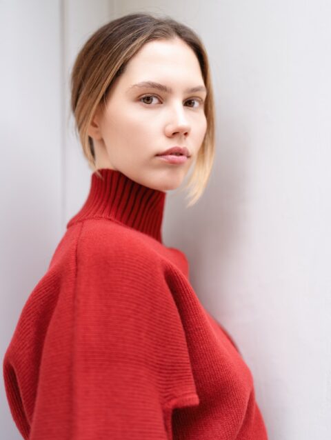 A woman in a red sweater leaning against a wall